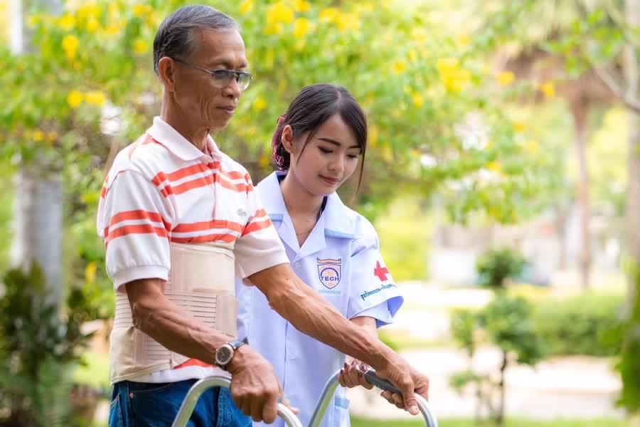 เจ้าหน้าที่ประคองผู้สูงอายุฝึกเดินด้วยวอล์คเกอร์ในสวนของศูนย์
